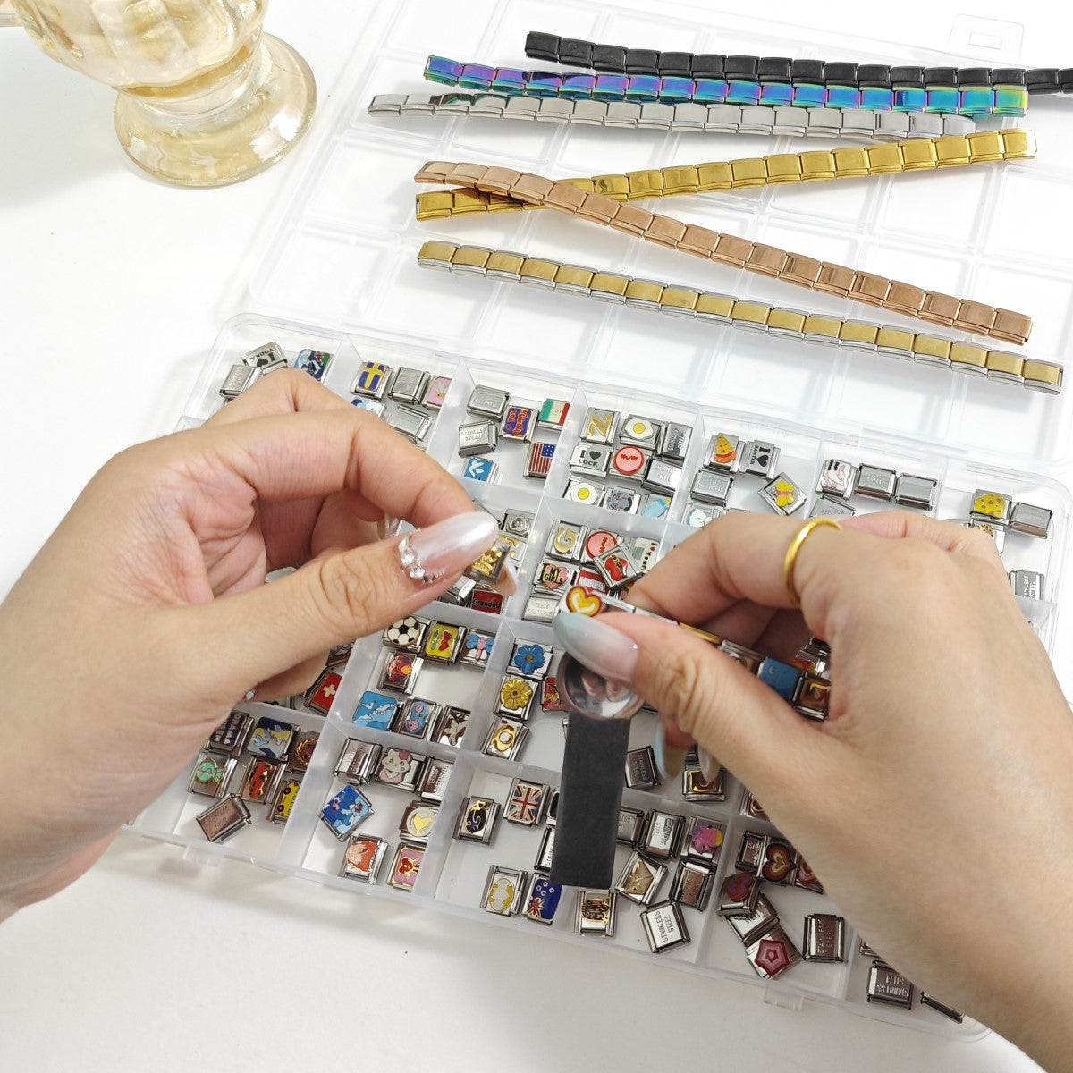 Hands using a charm tool to customize an Italian charm bracelet with assorted charm links laid out in an organizer