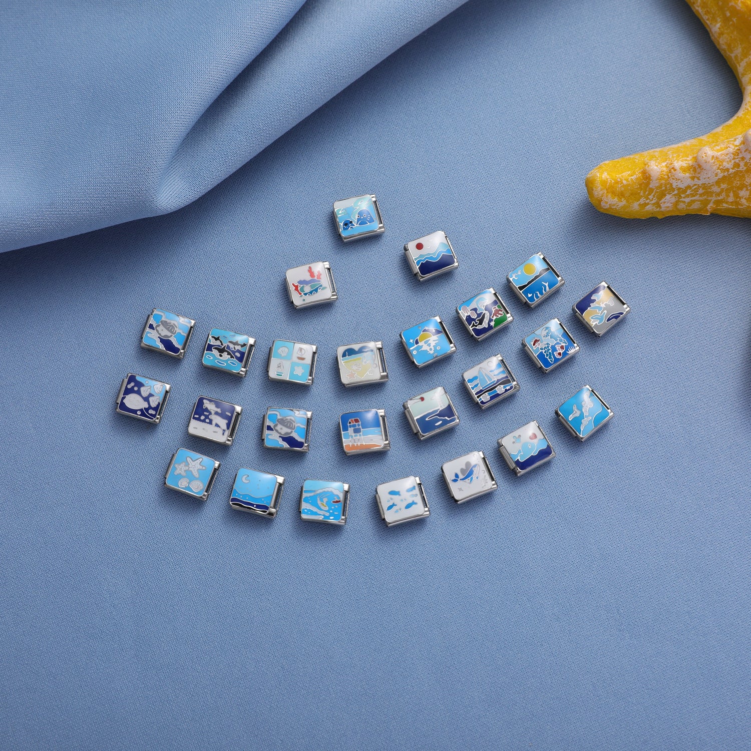 Ocean-themed Italian charm bracelet links with blue sea and coastal designs arranged on a soft blue fabric background