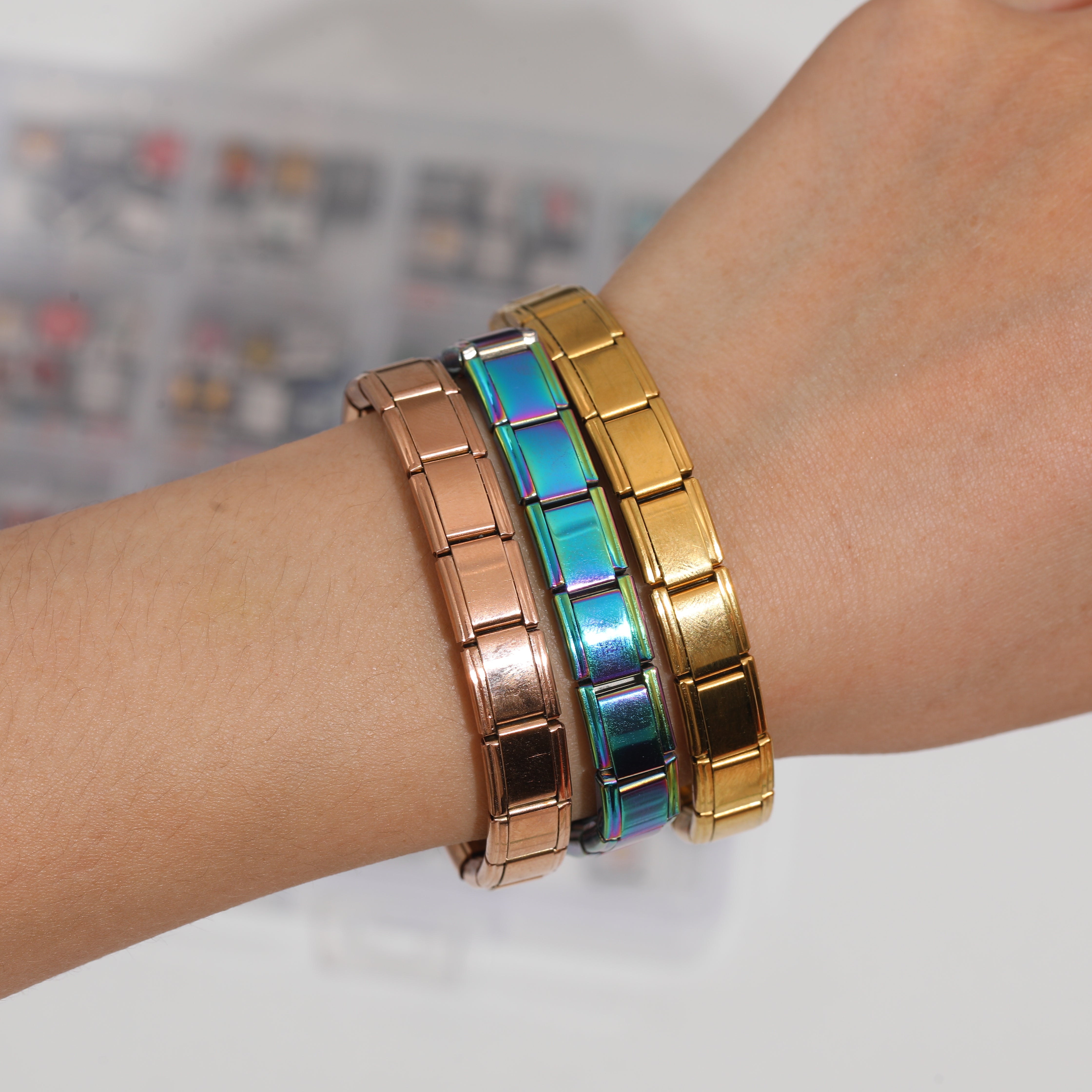 Close-up of a wrist wearing three stacked stainless steel Italian charm bracelets in iridescent rainbow rose gold and polished gold finishes against a plain white background