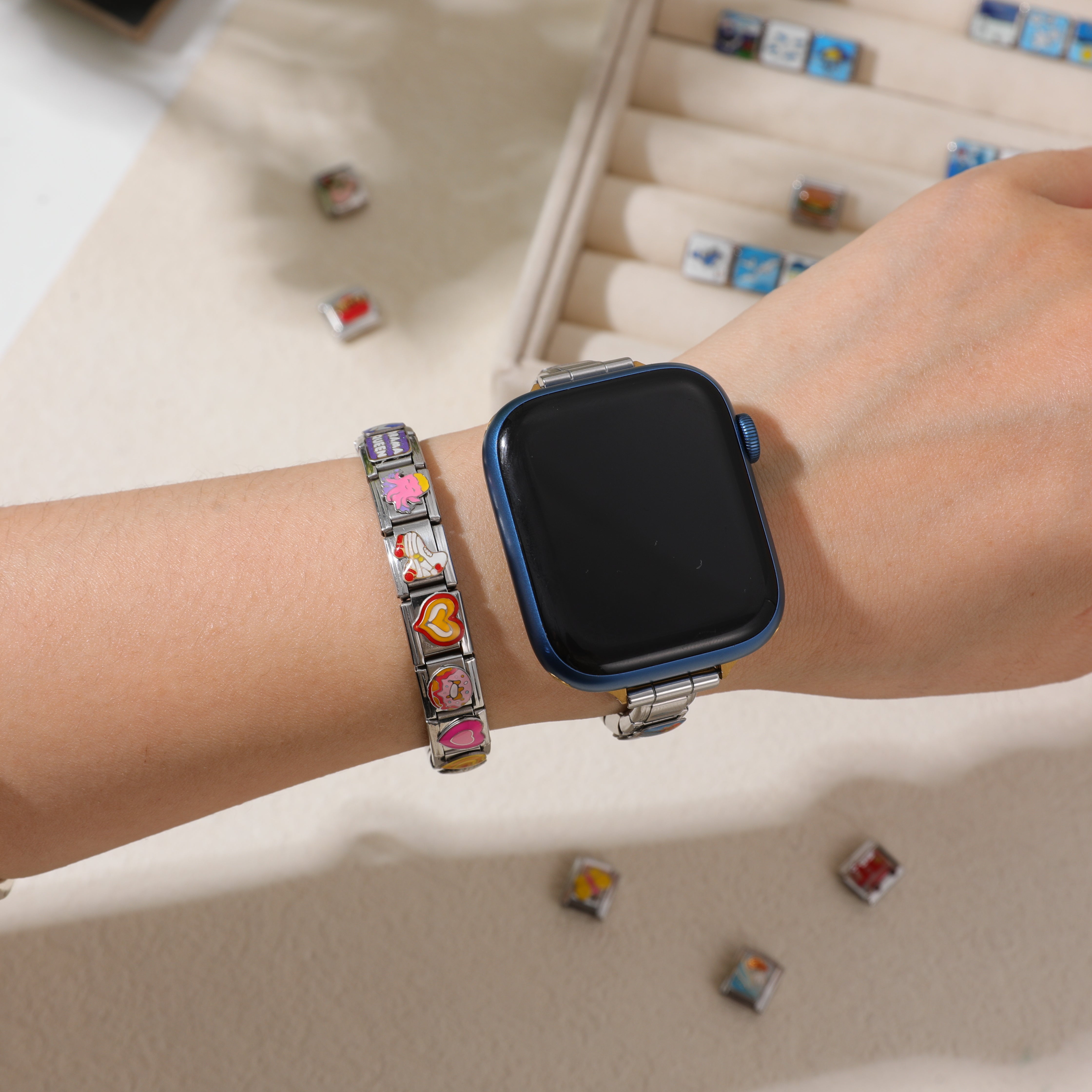 A wrist with a blue Apple Watch paired with a stainless steel Italian charm bracelet featuring colorful cartoon and heart-themed links