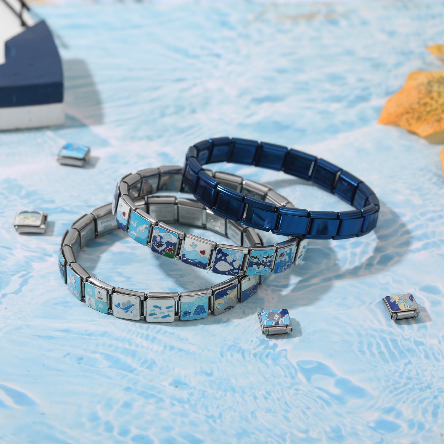 Stacked Italian charm bracelets with ocean-themed blue charms and a dark blue bracelet base on a water-pattern background
