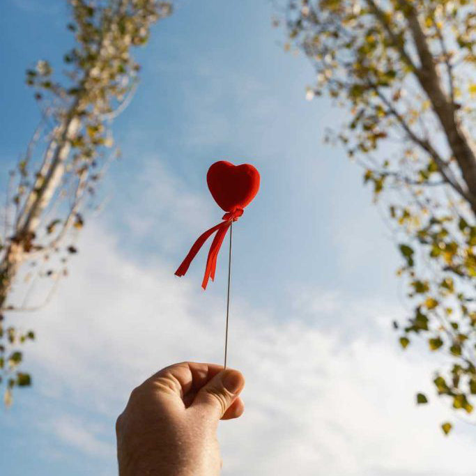 Hand holding a small red heart against the sky, symbolizing love and quiet affection